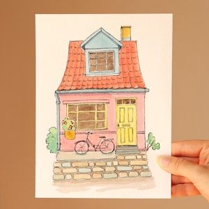 May include: A watercolour illustration of a pink house with a red tiled roof, a yellow door, and a dormer window. A bicycle is parked in front of the house, which sits on a stone foundation. Green bushes are visible on the sides of the structure.