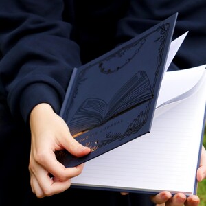 May include: A navy blue journal with a gold embossed design of an open book. The journal has lined pages and is open to reveal the blank pages.