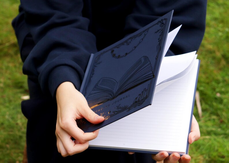 May include: A navy blue journal with a gold embossed design of an open book. The journal has lined pages and is open to reveal the blank pages.
