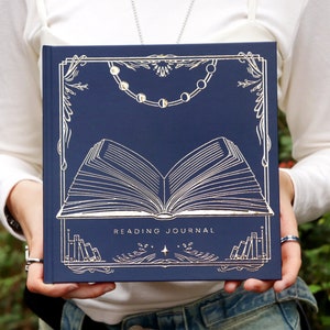 May include: A navy blue reading journal with a silver foil design of an open book, a crescent moon necklace, and the text "READING JOURNAL".