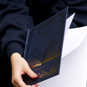 May include: A navy blue reading journal with gold foil accents. The cover features a stylized open book with the words "Reading Journal" printed on the bottom. The journal is open to a blank page with lined paper.