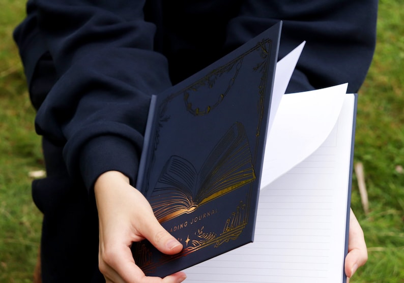 May include: A navy blue reading journal with gold foil accents. The cover features a stylized open book with the words "Reading Journal" printed on the bottom. The journal is open to a blank page with lined paper.