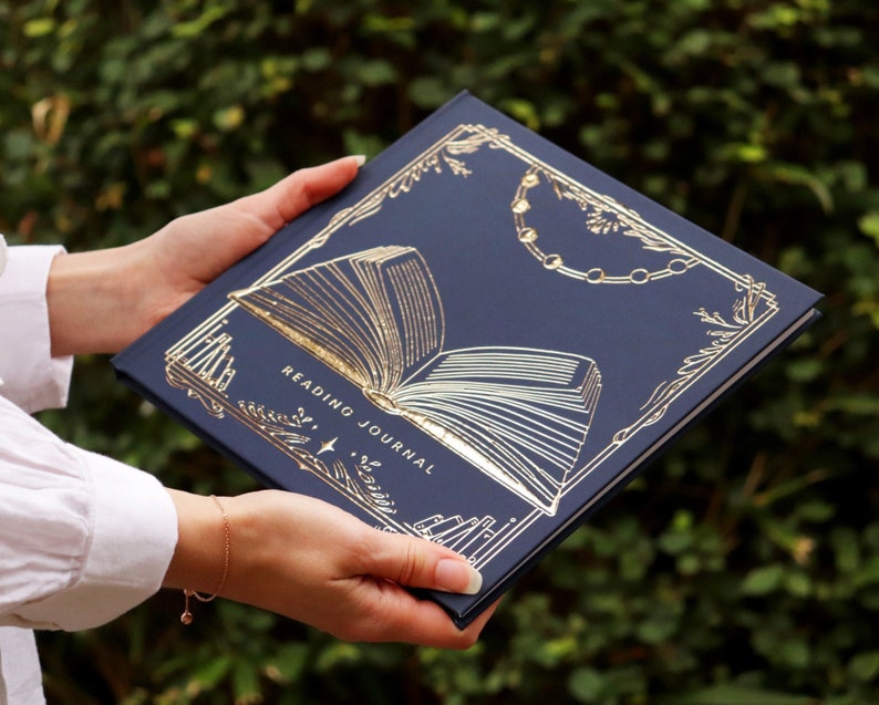 May include: A navy blue reading journal with a gold foil design of an open book and the words "Reading Journal" on the cover.