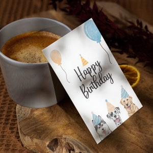 K&ouml;nnte beinhalten: Eine wei&szlig;e Karte mit dem Text "Happy Birthday" und drei Aquarell-Illustrationen von Hunden, die Partyh&uuml;te tragen. Die Karte liegt auf einer Holzoberfl&auml;che neben einer Tasse Kaffee.