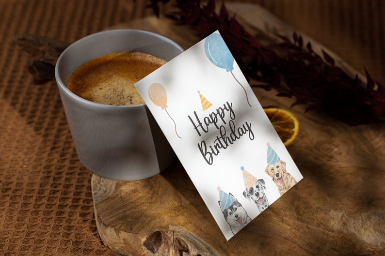 K&ouml;nnte beinhalten: Eine wei&szlig;e Karte mit dem Text "Happy Birthday" und drei Aquarell-Illustrationen von Hunden, die Partyh&uuml;te tragen. Die Karte liegt auf einer Holzoberfl&auml;che neben einer Tasse Kaffee.