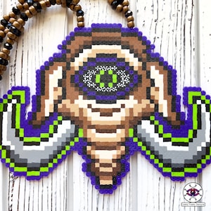 May include: A purple, green, brown, and white pixel art necklace featuring a creature with a single eye and large horns. The necklace is made of plastic beads and is strung on a brown cord.