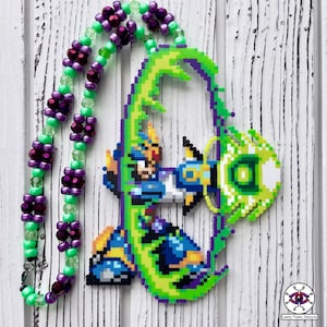 Pode incluir: Um colar de pixel art verde, azul e amarelo com um personagem de um videojogo. O personagem está segurando uma bola de energia verde. O colar é feito de contas e tem um esquema de cores roxo e verde.