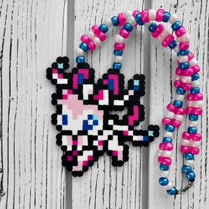 May include: A pink and blue kandi necklace with a pixelated image of a pink and blue creature. The necklace is made of small plastic beads.