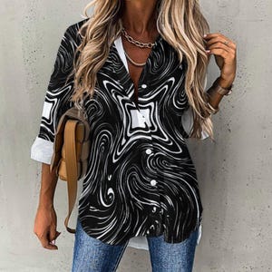 May include: A black and white patterned button-down shirt with a swirling design. The shirt has a V-neck, white cuffs, and a curved hemline. The sleeves are three-quarter length. The shirt is paired with blue jeans.