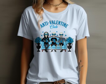 Könnte beinhalten: Hellblaues T-Shirt mit dem Schriftzug "ANTI-VALENTINE Club" in Blau. Die Grafik zeigt drei Skelettfiguren auf einem blauen Sofa, die Sonnenbrillen tragen und Tassen halten. Blaue Blumen befinden sich in Vasen auf beiden Seiten.
