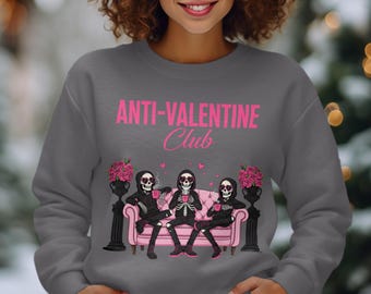 Könnte beinhalten: Graues Sweatshirt mit dem pinkfarbenen Text "ANTI-VALENTINE Club". Die Grafik zeigt drei Skelettfiguren auf einem rosa Sofa, jede mit einer Tasse. Schwarze Urnen mit rosa Blumen flankieren das Sofa.