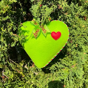 Peut inclure: Un ornement en forme de cœur vert lime vif avec une petite application de cœur en feutrine rouge sur le devant. L'ornement est surmonté d'un ruban vert clair imprimé du texte "Christmas" en rouge et bordé d'un cordon torsadé rouge, blanc et vert.