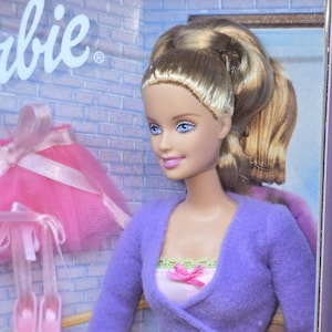 Puede incluir: Una muñeca Barbie con cabello rubio peinado en una coleta alta, ojos azules y una sonrisa rosa claro. La muñeca viste un cárdigan morado de manga larga sobre una blusa rosa claro con un pequeño lazo rosa. Se ve una falda de tul rosa y zapatillas.