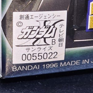 May include: A white rectangular sticker on a dark surface featuring black Japanese text, a copyright symbol, the letter B, and the number 0055022. Below the sticker, the text BANDAI 1996 MADE IN JAPAN is printed in white on a dark blue background.