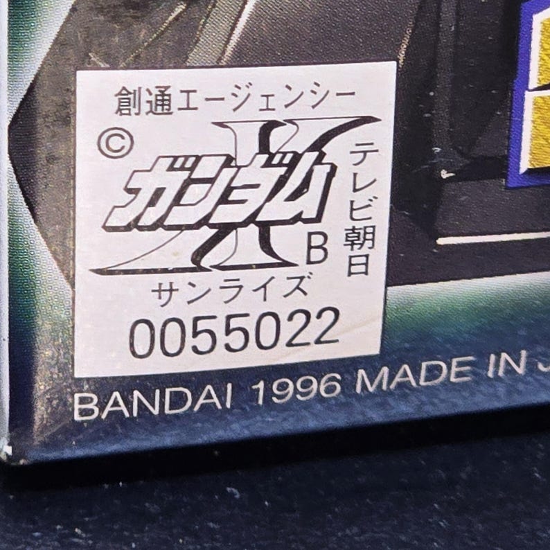 May include: A white rectangular sticker on a dark surface featuring black Japanese text, a copyright symbol, the letter B, and the number 0055022. Below the sticker, the text BANDAI 1996 MADE IN JAPAN is printed in white on a dark blue background.