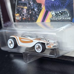 Puede incluir: Un coche Hot Wheels blanco y naranja con una cúpula transparente. El coche tiene neumáticos de banda blanca y detalles cromados en el motor. El embalaje incluye el texto "Hot Wheels Hall of Fame".