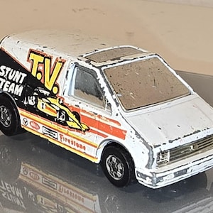 May include: White toy van with a red, yellow, and black "Stunt Team T.V." graphic on the side. The van has Firestone tires and is covered in dirt.