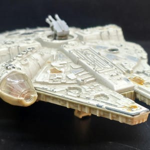 May include: A miniature off-white spaceship model featuring a circular main hull, a forward-facing dual-pronged nose, and a top-mounted turret. The surface is detailed with intricate mechanical paneling, recessed vents, and weathered metallic accents.