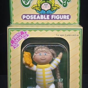 May include: A Cabbage Patch Kids poseable figure of Daria Jane in a yellow and white striped outfit, holding a small yellow bear. The doll is inside its original First Edition packaging, which features green and yellow accents and clear plastic.