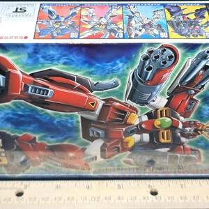May include: A rectangular cardboard box for a Gundam Leopard model kit featuring a red, white, and black robot illustration with a large cannon. A wooden ruler placed below the box shows the packaging is approximately 10 inches in length.