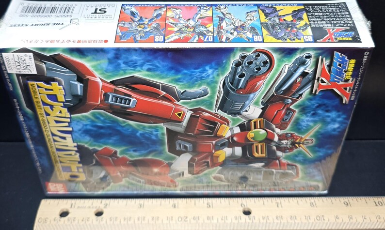 May include: A rectangular cardboard box for a Gundam Leopard model kit featuring a red, white, and black robot illustration with a large cannon. A wooden ruler placed below the box shows the packaging is approximately 10 inches in length.
