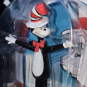 May include: A Cat in the Hat figurine encased in clear plastic packaging. The character features a tall red and white striped top hat, a red bow tie, white gloves, and a black and white body with a white chest patch and a long black tail.