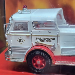 May include: A white and silver Mack fire engine model with red detailing. The vehicle displays the text "BALTIMORE FIRE DEPT." and the number "31". The wheels are black with red centres, and a red light is on top.