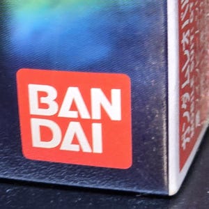 May include: A square red logo with the white text BAN DAI printed in two lines. The logo is positioned on the corner of a dark blue and white box. The text is bold and sans-serif, clearly visible against the solid red background of the square.