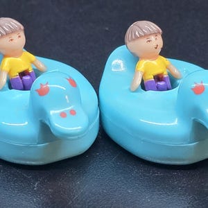 Può includere: Due piccoli giocattoli a forma di anatra azzurra, ciascuno con una figura in miniatura all'interno. Le figure hanno capelli castani, magliette gialle e pantaloni viola. Le anatre hanno dettagli rossi sulla testa.