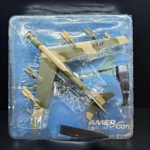 May include: A model airplane in a clear plastic package. The airplane is painted in camouflage colors of green, brown, and tan. The text "USAF" is visible on the wing. The package also includes the text "AMER COM COLLECTION".