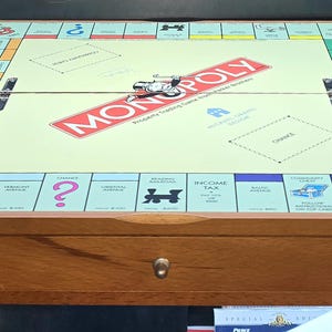 Op de afbeelding: Een houten Monopoly-bordspelset met een lichtgeel speeloppervlak, het klassieke rode Monopoly-logo, eigendomsvakken en spelbordafbeeldingen. Het bord is geïntegreerd in een houten basis met een lade aan de voorzijde en een knop.