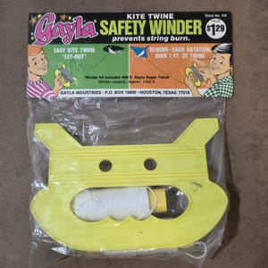 Op de afbeelding: Gele plastic drakenlijn oproller met een witte lijn erin. De oproller heeft twee gaten aan de bovenkant en een handvat aan de onderkant. De verpakking zegt "Gayla Safety Winder prevents string burn."