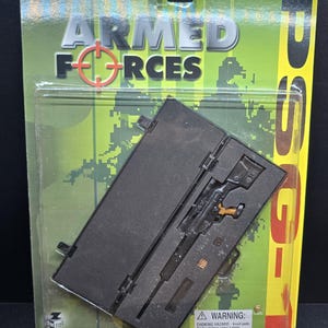 Puede incluir: Un rifle de francotirador de juguete en una funda negra, empaquetado con las palabras "ARMED FORCES" y una retícula de mira. El embalaje es verde y amarillo con una etiqueta de advertencia para mayores de 5 años. El rifle es negro con detalles dorados.