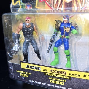 May include: A Judge Dredd action figure set featuring Lawgiver Dredd and an Under City Perp. The figures are in a clear plastic package with a cardboard backing. Dredd is in blue and green, while the Perp is in black and grey.