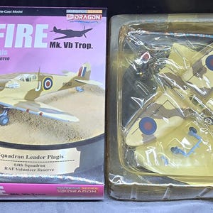 May include: A die-cast model of a Spitfire Mk. Vb Trop. aircraft, displayed on a base with a nameplate. The model is tan and brown with blue and red accents. The box reads "SPITFIRE" and "Squadron Leader Plagis".