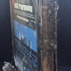 K&ouml;nnte beinhalten: Ein Commodore 64 Basic Programming Buch, noch in Plastik versiegelt. Das Cover zeigt einen blauen Himmel mit wei&szlig;en Strukturen. Das Buch ist von Peter Holmes & Derek Bush.