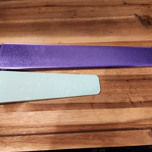 May include: Two rectangular desk accessories. One is a long, purple, textured item. The other is a smaller, light green item. Both are on a wooden surface. The items appear to be made of a solid material.