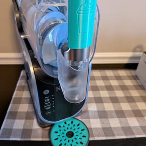 May include: A teal blue ice cream maker with a clear plastic container and a teal blue spout with a milkshake graphic. A teal blue round drip tray is in front of the machine.