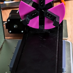 May include: A close-up of a pink and black 3D printer attachment. The attachment has three arms with black tips and a pink base. The attachment is mounted on a black metal track.