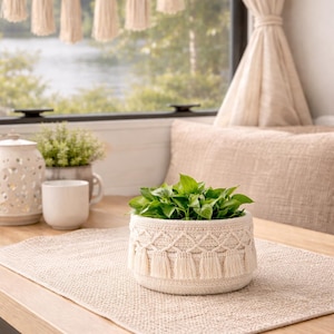 Könnte beinhalten: Ein gewebter beigefarbener Pflanzentopf mit Rautenmuster und Fransen, gefüllt mit grünen Pflanzen, steht auf einem passenden Platzset. Eine dekorative Quastengirlande hängt im Hintergrund und schafft eine gemütliche, natürliche Ästhetik.