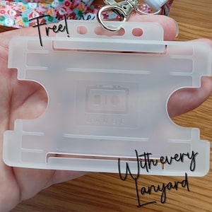 May include: A translucent, frosted plastic horizontal badge holder featuring a rectangular cutout design and a central embossed logo that reads BIO BADGE. The holder includes a top slot for a metal swivel hook attachment connected to a patterned lanyard.
