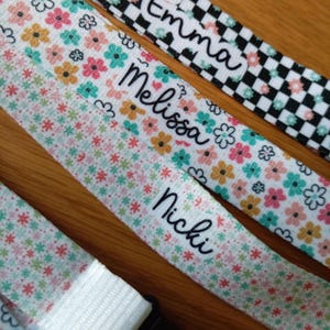May include: Three fabric wristbands featuring personalized names. One band has a black and white checkerboard pattern with the name Emma. Another features a floral print with the name Melissa. The third has a small floral pattern with the name Nicki.