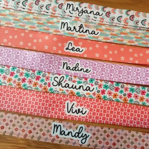 May include: A collection of seven patterned fabric ribbons, each featuring a unique design including rainbows, stripes, flowers, hearts, and peace signs. Each ribbon displays a white label with a black name: Mirjana, Martina, Lea, Nadine, Shauna, Vivi, and Mandy.