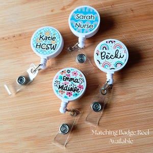 May include: Four circular white badge reels with retractable clear plastic straps and metal clips. Each reel features a unique colorful design: one with yellow flowers, one with blue rainbows, one with multicolored rainbows, and one with pink and orange flowers.