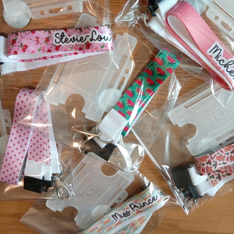 May include: A collection of fabric lanyards with various patterns including cherries, watermelons, polka dots, and gingerbread men, each featuring a clear plastic ID badge holder, a metal lobster clasp, and personalized names like Stevie-Lou and Michelle.
