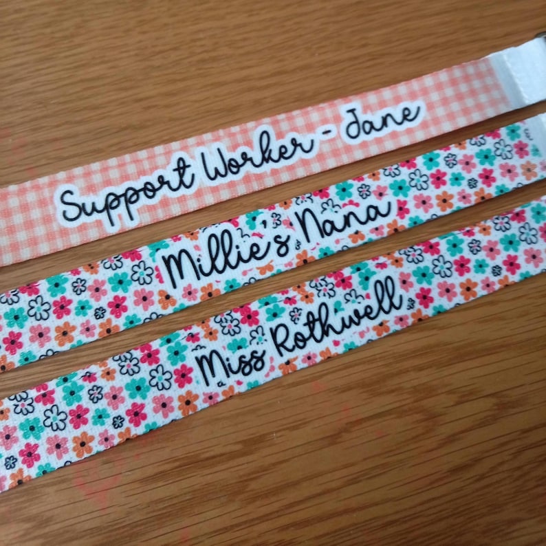 May include: Three fabric lanyards with black cursive text. The top lanyard has a peach gingham pattern and reads Support Worker - Jane. The middle and bottom lanyards feature a multi-colored floral print and read Millie's Nana and Miss Rothwell.