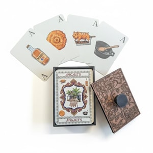 May include: A deck of playing cards with the word "ANGALE'S" on the box. The cards feature illustrations of food and drink items. The deck is next to a wooden card holder with a black knob.