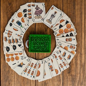 May include: A deck of playing cards arranged in a circular pattern around a green rectangular box with the text Boricua Travel Cards embossed on the lid. The cards feature various illustrations including figures, food items, and bottles in a colorful style.