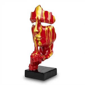Op de afbeelding: Een rode en gouden abstracte sculptuur van een gezicht met een vinger op de lippen, op een zwarte basis. De sculptuur heeft gouden verf die over het rode gezicht druipt. De basis is een zwarte vierkant.