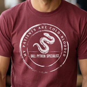 May include: A maroon t-shirt with a white circular graphic featuring a snake and the text "MY PATIENTS ARE COLD-BLOODED BALL PYTHON SPECIALIST". The shirt is made of a soft material.